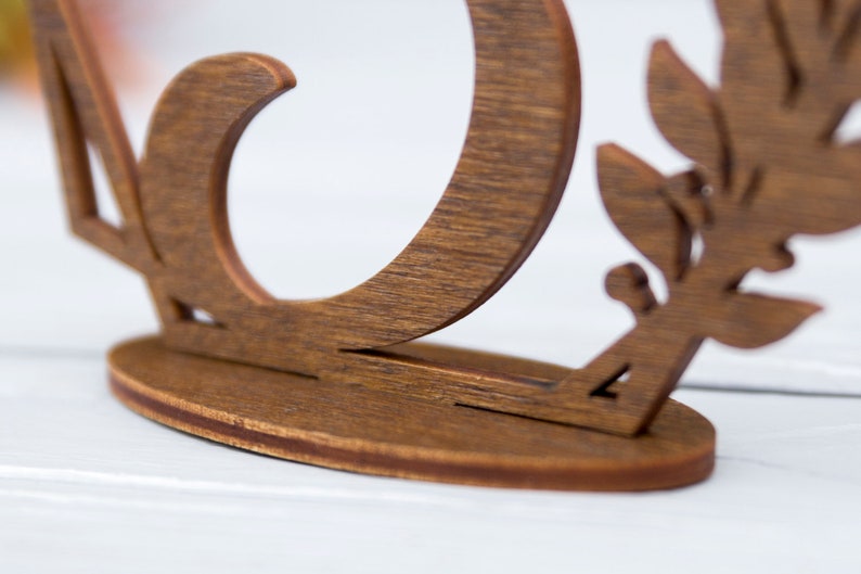 May include: A wooden table number holder with a decorative leaf design. The number is cut out of the wood and the holder has a rounded base.