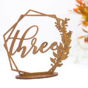 May include: Wooden table number with a geometric frame and floral design. The number "Three" is written in cursive script.