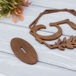 May include: Wooden table number with the number 51 cut out in the center. The number is surrounded by a geometric frame and leafy details. The table number is on a wooden stand.