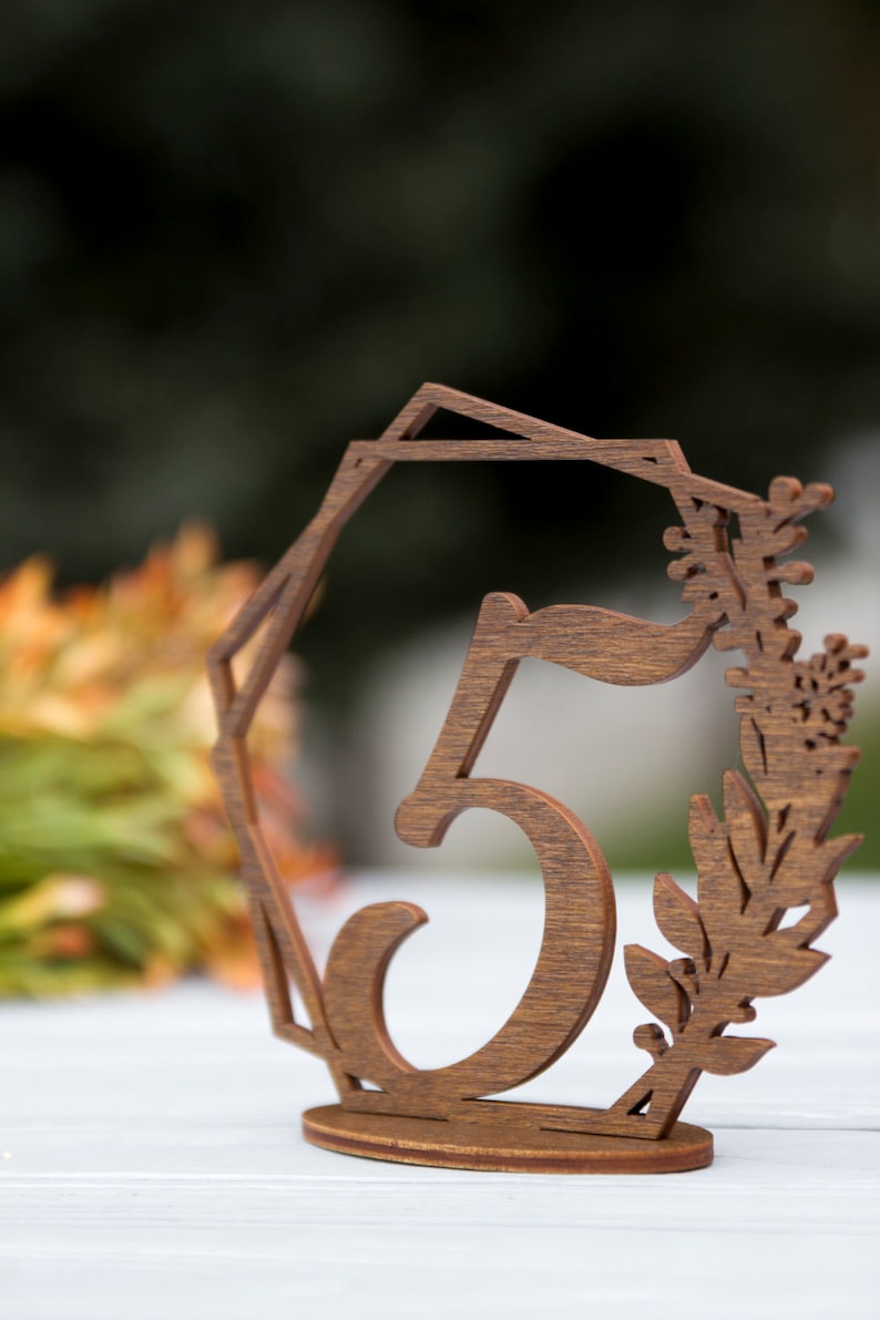 May include: Wooden table number with the number 5 cut out in the center. The number is surrounded by a decorative floral design.