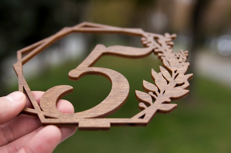 May include: A wooden table number with the number "5" cut out in the center. The number is surrounded by a decorative floral design.