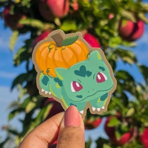 May include: A sticker featuring a green cartoon character with a pumpkin on its back. The pumpkin is orange with a green stem. The character has red eyes and white teeth. The sticker is held in front of a background of red apples and green leaves.