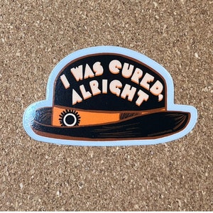 May include: A black and orange bowler hat sticker with the text "I was cured, alright".