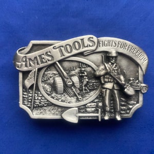 May include: A silver belt buckle with a raised design of a soldier holding a shovel and a cannon. The buckle has the text "Ames Tools Fights For Freedom" on it.