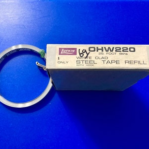 May include: A Lufkin OHW220 25 foot 8ths white clad steel tape refill with hook. The tape measure is rolled up and the box is white with black text.