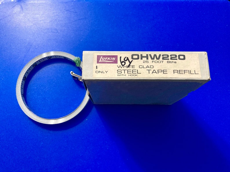 May include: A Lufkin OHW220 25 foot 8ths white clad steel tape refill with hook. The tape measure is rolled up and the box is white with black text.