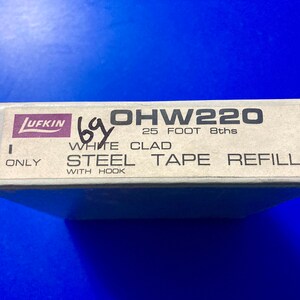 May include: A white box with black text that reads "OHW220 25 FOOT 8ths WHITE CLAD STEEL TAPE REFILL WITH HOOK ONLY". The box has a purple and white label that reads "LUFKIN".