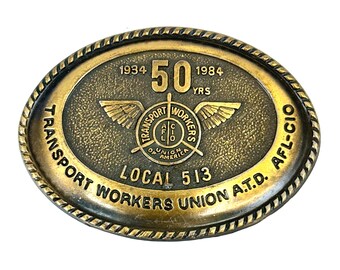 May include: An oval brass belt buckle with a rope-style border. The center features the text 1934 50 YRS 1984, a winged emblem with the text TRANSPORT WORKERS UNION OF AMERICA AFL-CIO, and the text LOCAL 513 TRANSPORT WORKERS UNION A.T.D. AFL-CIO.
