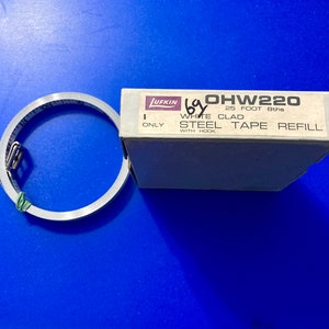May include: A white box with the text "OHW220 25 FOOT 8ths WHITE CLAD STEEL TAPE REFILL WITH HOOK ONLY" on it. A silver metal tape measure is next to the box.