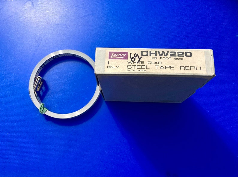 May include: A white box with the text "OHW220 25 FOOT 8ths WHITE CLAD STEEL TAPE REFILL WITH HOOK ONLY" on it. A silver metal tape measure is next to the box.