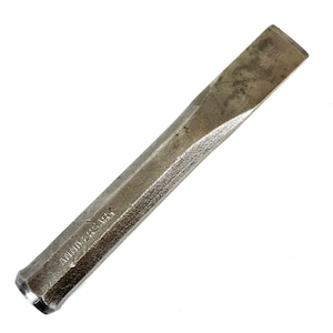 May include: A silver-toned metal cold chisel with a hexagonal shaft and a beveled cutting edge. The word ANNIVERSARY is stamped into the side of the metal tool. The surface shows a textured, weathered finish with signs of use and minor oxidation.