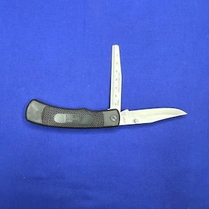 May include: A black folding pocket knife with a silver blade and a ruler on the back. The knife has a black handle with the word "CRAFT" printed on it. The blade is partially open and the ruler is visible. The knife is on a blue background.