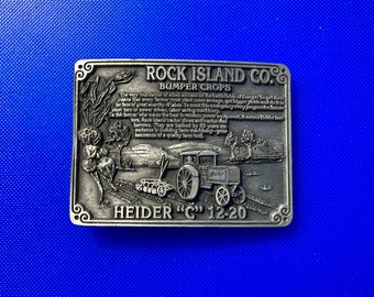 May include: A silver-toned metal belt buckle with an image of a tractor and the text "Rock Island Co. Bumper Crops Heider "C" 12-20".