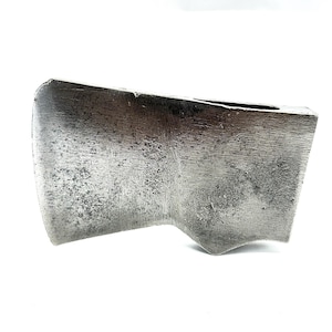 May include: Close-up of a vintage axe head. The metal head has a worn, textured appearance with a curved blade and a rectangular top. The axe head is a dull silver color, suggesting age and use.