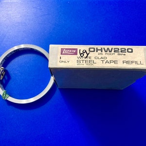 May include: A white box with the text "OHW220 25 FOOT 8ths WHITE CLAD STEEL TAPE REFILL WITH HOOK ONLY" printed on it. The box contains a metal tape measure.