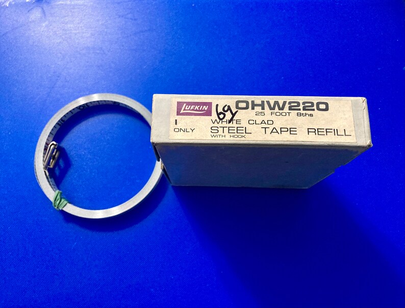 May include: A white box with the text "OHW220 25 FOOT 8ths WHITE CLAD STEEL TAPE REFILL WITH HOOK ONLY" printed on it. The box contains a metal tape measure.