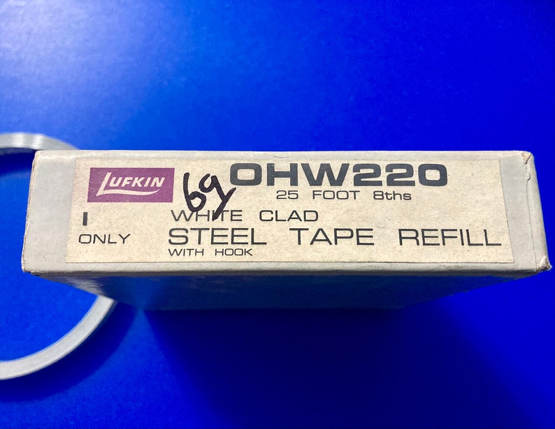 May include: A white box with the text "Lufkin OHW220 25 FOOT 8ths WHITE CLAD STEEL TAPE REFILL ONLY WITH HOOK" printed on it.