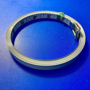 May include: A white measuring tape with black markings is coiled into a circle on a blue surface. The tape is 6 inches long and has a metal clasp.