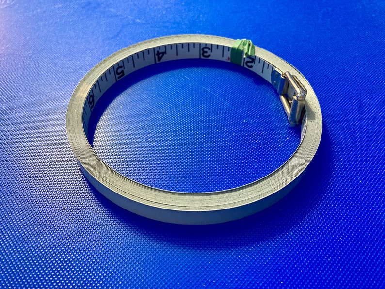 May include: A white measuring tape with black markings is coiled into a circle on a blue surface. The tape is 6 inches long and has a metal clasp.