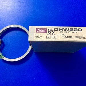 May include: A white box with the text "OHW220 25 FOOT 8ths WHITE CLAD STEEL TAPE REFILL WITH HOOK" on it. A rolled up white tape measure is next to the box.