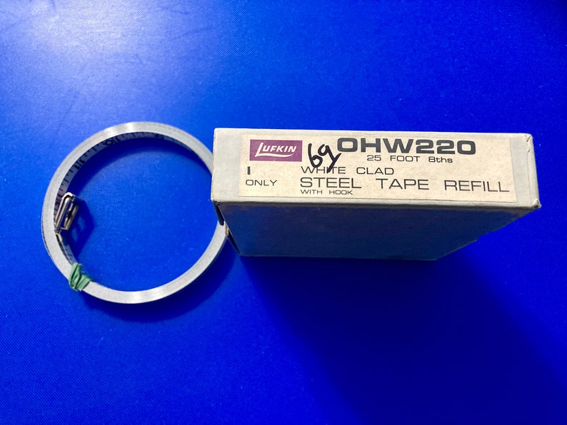 May include: A white box with the text "OHW220 25 FOOT 8ths WHITE CLAD STEEL TAPE REFILL WITH HOOK" on it. A rolled up white tape measure is next to the box.