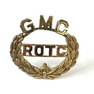 May include: A gold-toned ROTC pin with the letters GMC at the top and ROTC in the center. The pin is surrounded by a laurel wreath design and has a small decorative element at the bottom.