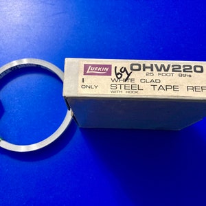 May include: A white box with the text "OHW220 25 FOOT 8ths WHITE CLAD STEEL TAPE REFILL WITH HOOK" and the Lufkin logo. A silver tape measure is next to the box.