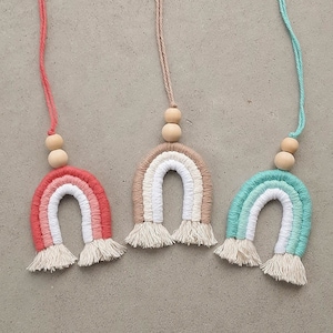 Może przedstawiać: Trzy makramowe ozdoby w kształcie tęczy w kolorach koralowym, beżowym i turkusowym. Każda tęcza ma biały środek, frędzelkową podstawę i drewniane koraliki. Wiszą na pasujących kolorystycznie sznurkach.
