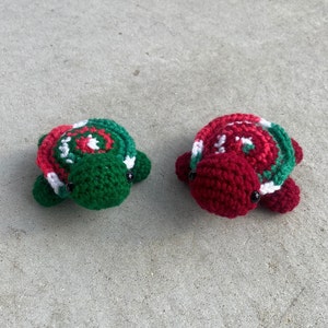 May include: Two handmade crochet turtles. One is green with red and white accents, the other is red with green and white accents. Both have black eyes and are lying on a concrete surface. These decorative turtles are perfect for a nursery or as a gift.