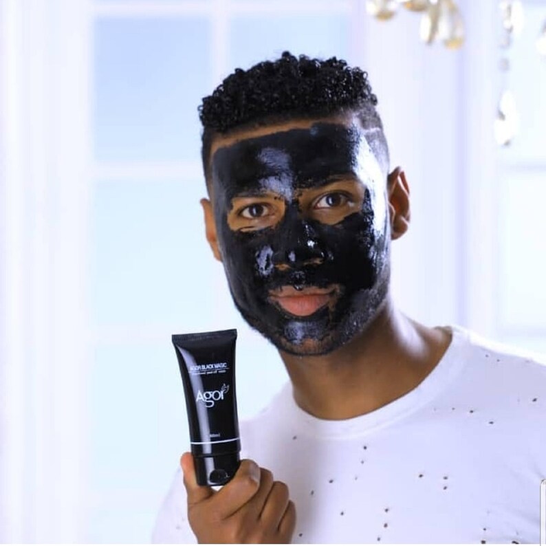 May include: A man wearing a black charcoal face mask holds a tube of black face mask product labeled "Deep Black Mask" with the brand name "Agor" printed on the tube.