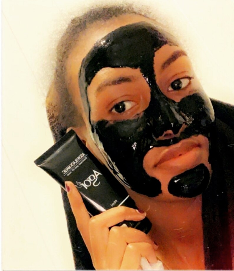 May include: A person is applying a black charcoal face mask. The mask is covering most of their face, leaving only their eyes and mouth visible. They are holding a black tube with the text "AOGA BLACK MASK" printed on it.