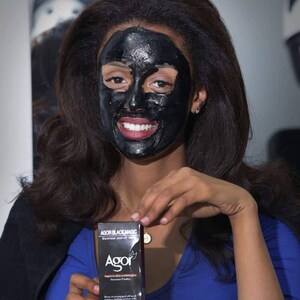 May include: A woman with long black hair is wearing a black face mask. She is holding a black box with the text "AGOR BLACK MAGIC" and "AGOR" on it. The box is a blackhead peel off mask.