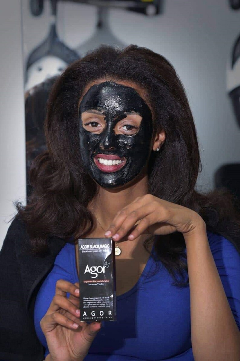 May include: A woman with long black hair is wearing a black face mask. She is holding a black box with the text "AGOR BLACK MAGIC" and "AGOR" on it. The box is a blackhead peel off mask.