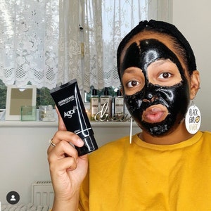 May include: A person wearing a black face mask and a yellow shirt is holding a black tube of face mask product labeled "AGOR Black Magic Activated Charcoal Peel Off Mask 60ml".