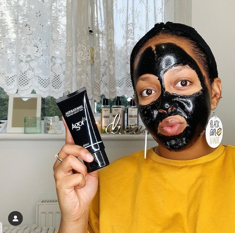May include: A person wearing a black face mask and a yellow shirt is holding a black tube of face mask product labeled "AGOR Black Magic Activated Charcoal Peel Off Mask 60ml".