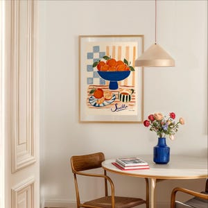 K&ouml;nnte beinhalten: Ein gerahmter Kunstdruck mit einem Stillleben von Orangen in einer blauen Schale und auf einem Teller, mit dem Wort "Sevilla" darunter. Der Druck befindet sich in einem Raum mit einem runden Tisch, einer blauen Blumenvase und einer Pendelleuchte.