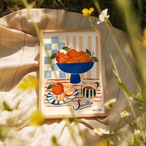 K&ouml;nnte beinhalten: Ein gerahmter Kunstdruck mit einem Stillleben mit Orangen. Eine blaue Sch&uuml;ssel mit Orangen steht auf einem Tisch mit gestreiftem Hintergrund. Ein Teller mit geschnittenen Orangen und eine Tasse befinden sich ebenfalls auf dem Tisch. Das Wort "Sevilla" ist kursiv geschrieben.