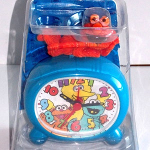 May include: A blue alarm clock with Elmo's face on the front. The clock features Sesame Street characters Big Bird, Cookie Monster, and Elmo. The clock is packaged in a clear plastic clamshell. The text on the packaging reads "Elmo's Quartz Table Alarm Clock".