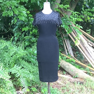 May include: A black sleeveless dress with a beaded neckline and a fitted silhouette. The dress is displayed on a mannequin against a backdrop of green foliage.