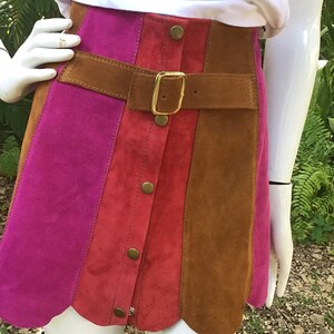 May include: A suede mini skirt with a scalloped hem. The skirt is made of three panels of different colours: pink, red, and brown. The skirt has a brown leather belt with a gold buckle and a row of gold buttons down the front.