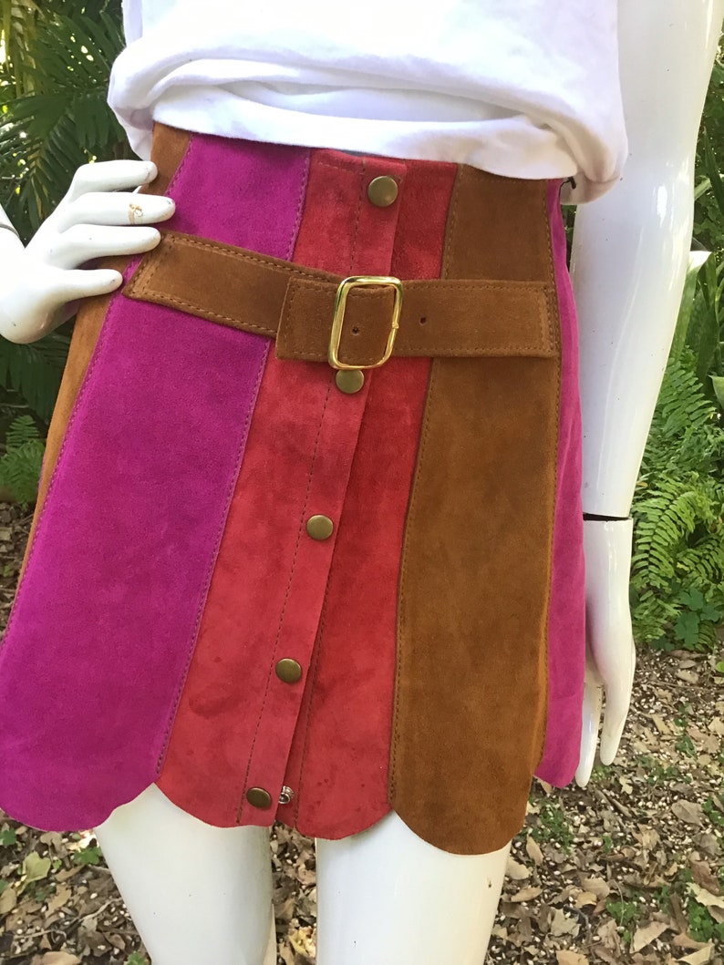May include: A suede mini skirt with a scalloped hem. The skirt is made of three panels of different colours: pink, red, and brown. The skirt has a brown leather belt with a gold buckle and a row of gold buttons down the front.