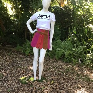 May include: A white t-shirt with a black heart and the text "PLAY" and "COMME des GARCONS" on the front. The mannequin is wearing a multi-coloured mini skirt with a brown leather belt.