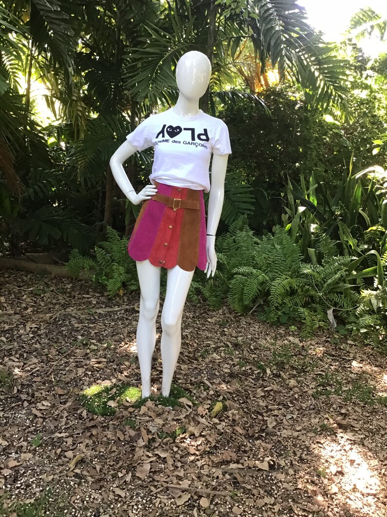 May include: A white t-shirt with a black heart and the text "PLAY" and "COMME des GARCONS" on the front. The mannequin is wearing a multi-coloured mini skirt with a brown leather belt.