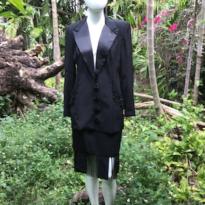 May include: A black blazer with a satin lapel and a matching skirt. The skirt has a fringe hem.