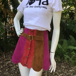 May include: A pink, red and brown suede mini skirt with a scalloped hem and a gold buckle belt. The skirt has a button closure and is worn with a white t-shirt that says "Play" with a black heart and "Comme des Gar&ccedil;ons" underneath.
