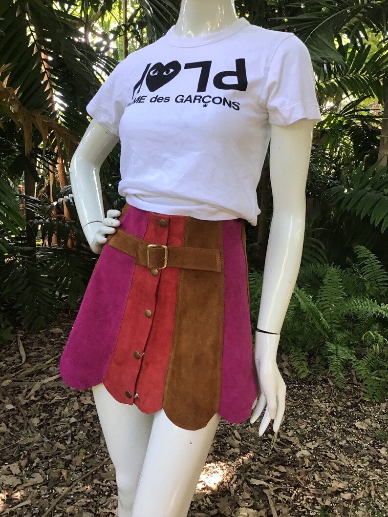 May include: A pink, red and brown suede mini skirt with a scalloped hem and a gold buckle belt. The skirt has a button closure and is worn with a white t-shirt that says "Play" with a black heart and "Comme des Gar&ccedil;ons" underneath.