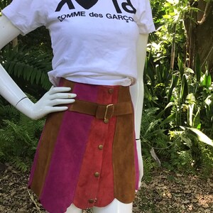May include: A brown, pink and red suede mini skirt with a scalloped hem. The skirt has a brown leather belt with a gold buckle and a row of gold buttons down the front.