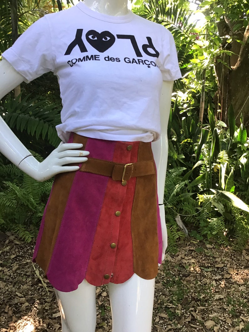 May include: A brown, pink and red suede mini skirt with a scalloped hem. The skirt has a brown leather belt with a gold buckle and a row of gold buttons down the front.
