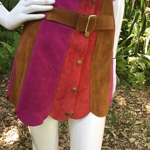 May include: A suede mini skirt with a brown leather belt and gold buckle. The skirt is made of three panels of different colours: pink, red, and brown. The skirt has a scalloped hem.