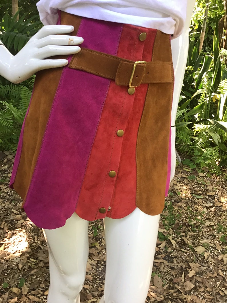 May include: A suede mini skirt with a brown leather belt and gold buckle. The skirt is made of three panels of different colours: pink, red, and brown. The skirt has a scalloped hem.